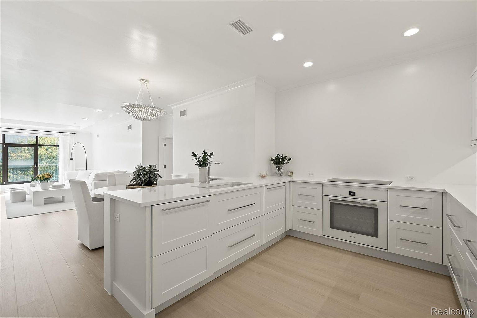 This is a bright, modern kitchen with white cabinetry, countertops, and walls, creating a clean and spacious feel. The kitchen features a central island with a sink, stainless steel appliances including a built-in oven, and light wood flooring. The open-concept design seamlessly connects the kitchen to the living area, enhancing the sense of flow and connectivity within the home.