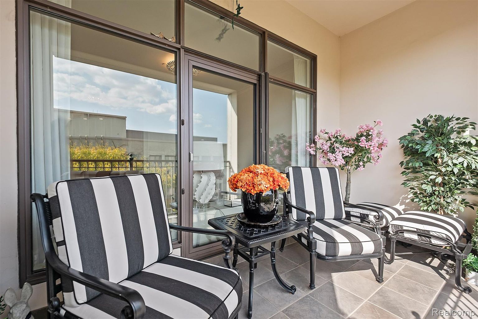 This image showcases a stylish balcony setup, featuring two black and white striped chairs and a small table adorned with a vibrant orange floral arrangement. The balcony is enclosed by large windows, offering a glimpse of the outside scenery. The overall aesthetic is modern and inviting, perfect for relaxation and enjoying the view.