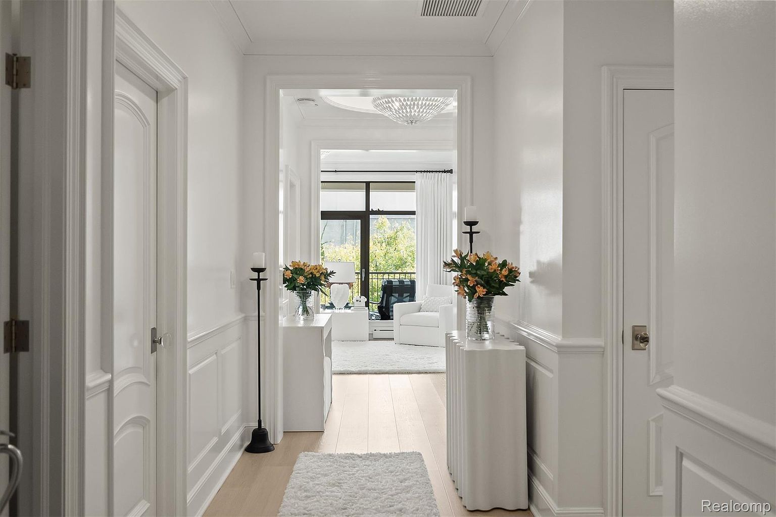 This is an interior shot of a bright, elegant hallway. The hallway is painted white and features detailed trim work and paneling. A doorway at the end of the hall leads into a living area with a view of the outdoors, creating a sense of depth and openness.