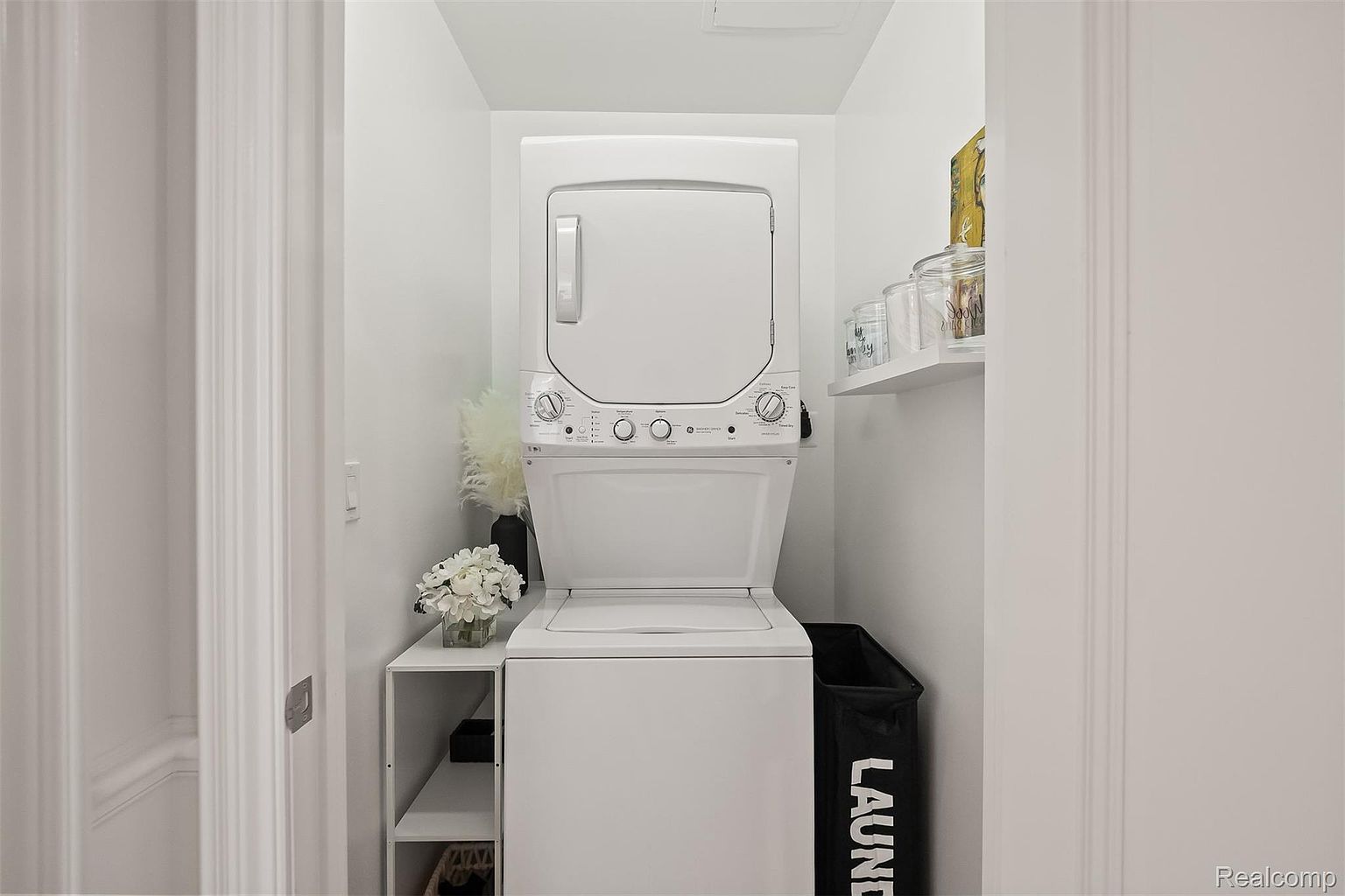This is a well-organized laundry room featuring a stacked washer and dryer unit. A small shelving unit to the left provides storage, while a laundry hamper sits to the right. The room is painted in a clean white, creating a bright and functional space.