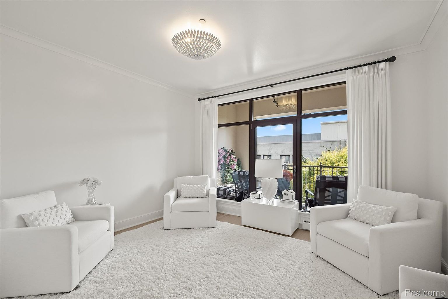 The image showcases a bright and airy living room featuring three white armchairs arranged around a plush white rug. A modern chandelier illuminates the space, while a large window with white curtains provides natural light and a view of the outdoors. The room exudes a clean, minimalist aesthetic with a focus on comfort and elegance.