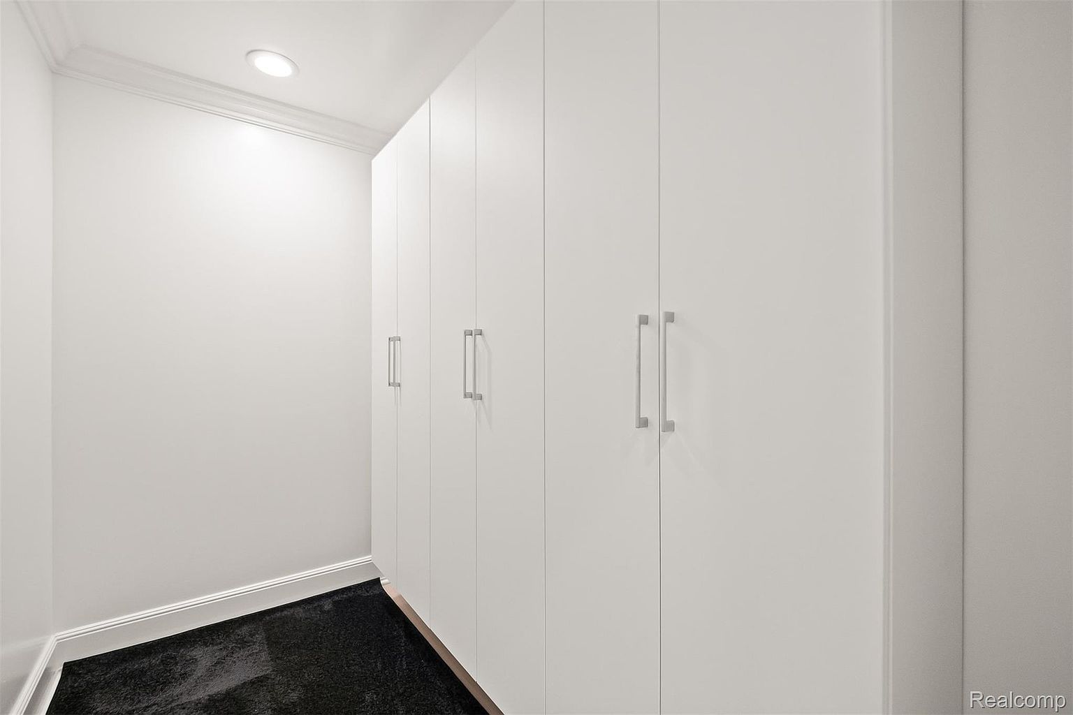 This is an interior shot of a walk-in closet featuring a series of white, floor-to-ceiling cabinets with sleek, silver handles. The closet has dark carpeting and is well-lit by a recessed ceiling light. The overall impression is clean, modern, and organized, suggesting ample storage space.