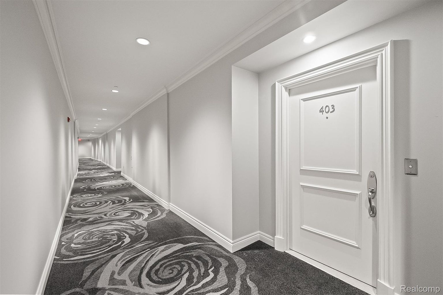 This is an interior shot of a well-lit hallway in an apartment building or condominium. The hallway features a patterned carpet with a floral design, white walls, and recessed lighting. A door with the number '403' is visible on the right, suggesting an apartment unit.