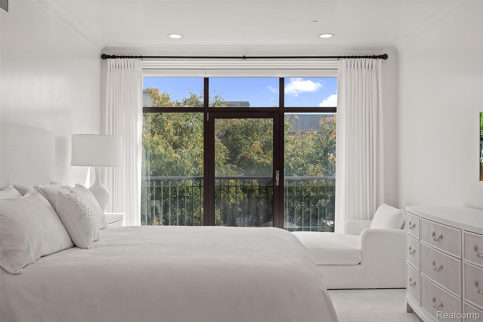 This is a bright and airy primary bedroom featuring a large window with a balcony overlooking lush greenery. The room is decorated in a minimalist style with white bedding, curtains, and furniture, creating a serene and inviting atmosphere. A dresser with decorative knobs adds a touch of elegance.