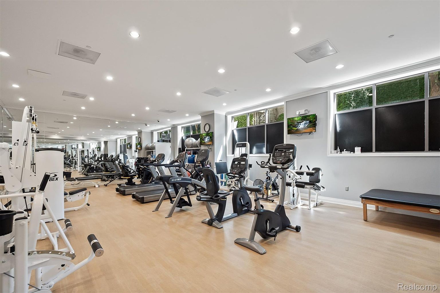 This is a spacious home gym featuring a variety of exercise equipment, including treadmills, stationary bikes, and weight machines. The room has light wood flooring, light gray walls, and large windows that provide natural light. A mirrored wall enhances the sense of space, and the overall impression is clean and well-equipped.