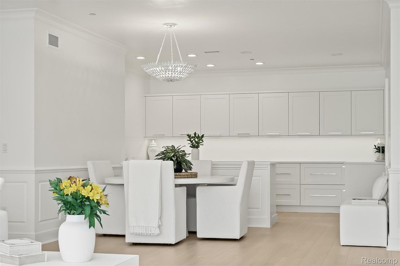 This is a bright and airy dining room featuring a round white table surrounded by white upholstered chairs. A modern chandelier hangs above the table, and built-in white cabinets provide ample storage. The room has a clean, minimalist aesthetic with light wood flooring and white walls, creating a sophisticated and inviting space.