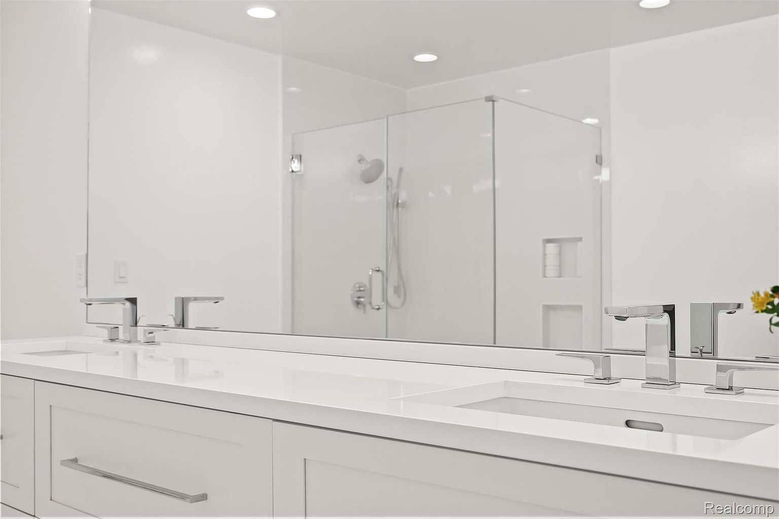 This is a bright and modern primary bathroom featuring a double vanity with white cabinetry and a white countertop. The faucets are sleek and chrome, and a large mirror reflects a glass-enclosed shower with chrome fixtures. The overall impression is clean, minimalist, and luxurious.