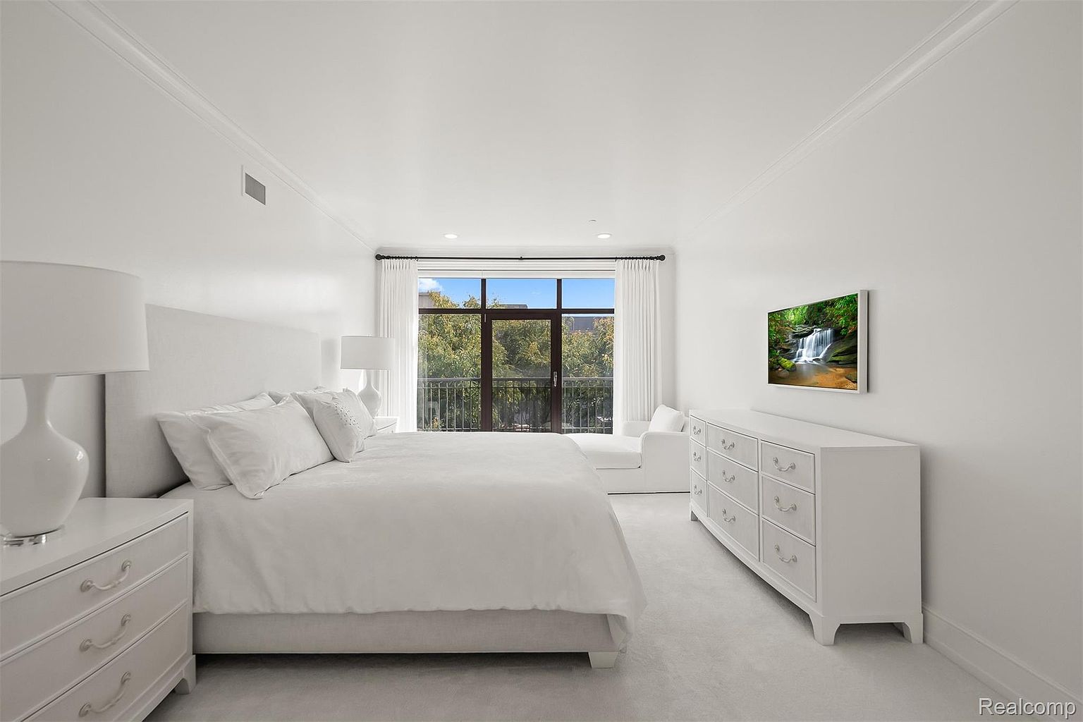This is a bright and airy primary bedroom featuring an all-white color scheme. The room includes a large bed with crisp white linens, a matching dresser with a television above it, and a balcony with a view of trees. The overall impression is clean, modern, and serene, creating a relaxing atmosphere.