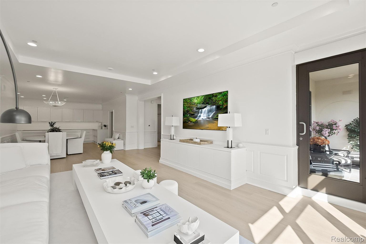 This is a bright and airy living room featuring a modern aesthetic with white walls, light wood flooring, and minimalist decor. A large flat-screen TV is mounted on the wall above a white console table, flanked by matching lamps. The room is open to the kitchen and dining area, creating a spacious and inviting atmosphere.
