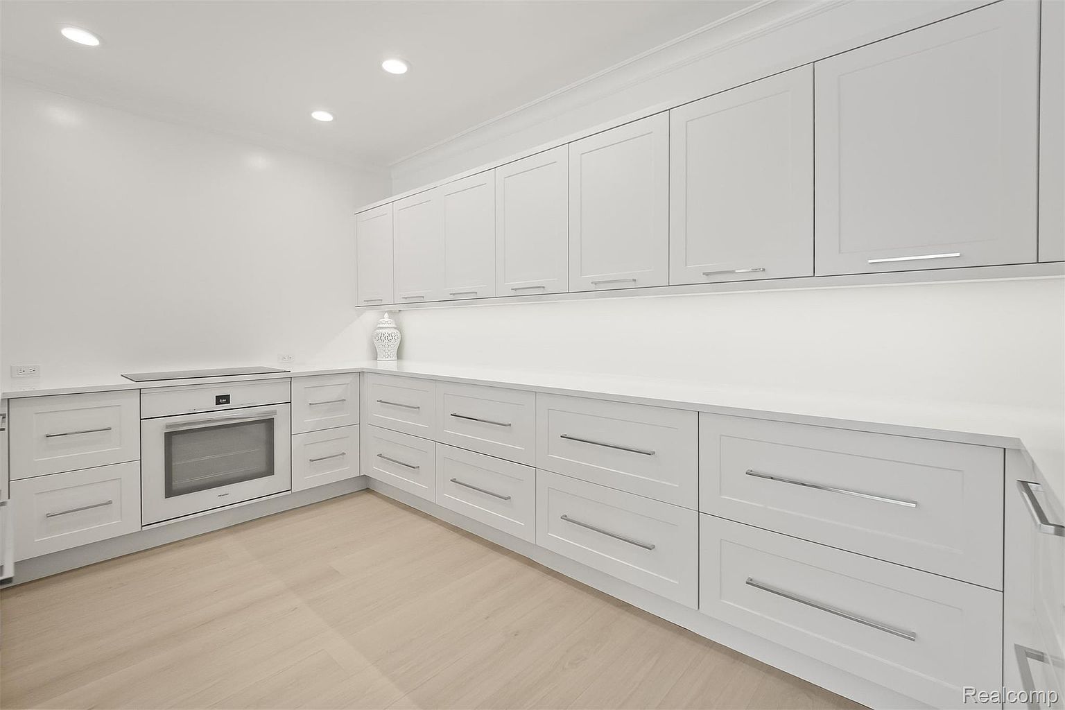 This is a bright, modern kitchen featuring sleek white cabinetry with silver hardware and light wood flooring. The kitchen is well-lit with recessed lighting and under-cabinet lighting, highlighting the clean countertops and built-in oven. The overall impression is one of minimalist elegance and functionality.