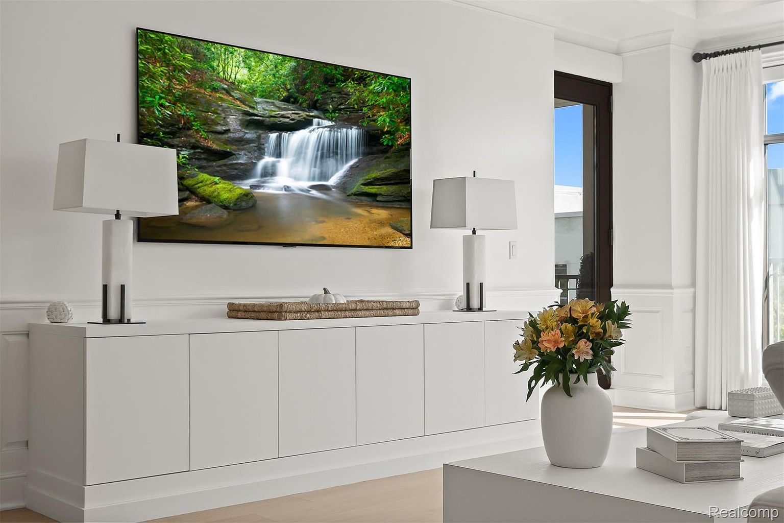 This is a bright and airy living room featuring a large flat-screen TV displaying a waterfall scene, mounted above a long, white cabinet. Two modern lamps flank the TV, adding to the room's sophisticated ambiance. A vase of flowers sits on a coffee table in the foreground, adding a touch of color and life to the space.