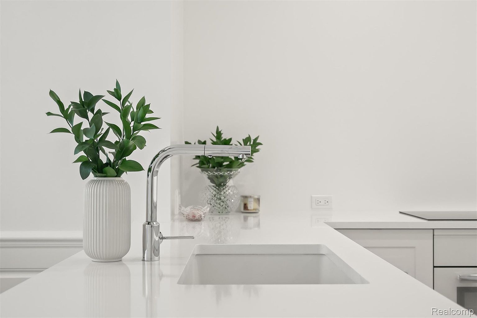 A bright, modern kitchen features a clean white countertop with a stainless steel faucet and a deep sink. A decorative vase with greenery adds a touch of nature, while another smaller plant arrangement sits in the background. The overall impression is one of minimalist elegance and functionality.