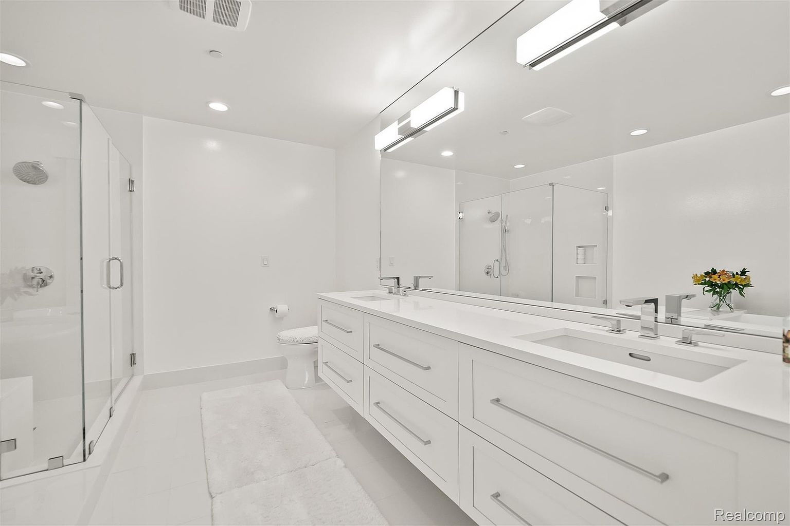 This is a bright and modern primary bathroom featuring a double vanity with sleek white cabinetry and countertops, complemented by contemporary chrome fixtures. A large mirror spans the length of the vanity, reflecting the clean lines of the glass-enclosed shower. The all-white color scheme creates a spacious and luxurious feel.
