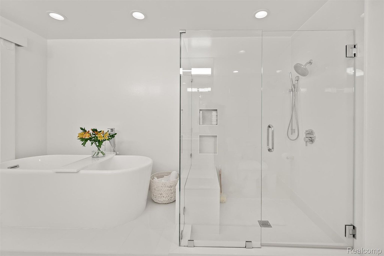 This is a bright and modern primary bathroom featuring a freestanding white bathtub with a small bouquet of flowers, a glass-enclosed shower with a rainfall showerhead and built-in shelving, and a white vanity. The bathroom has a clean and minimalist design with white walls and tile flooring, creating a spa-like atmosphere. The perspective is from the doorway, showcasing the layout and key features of the bathroom.
