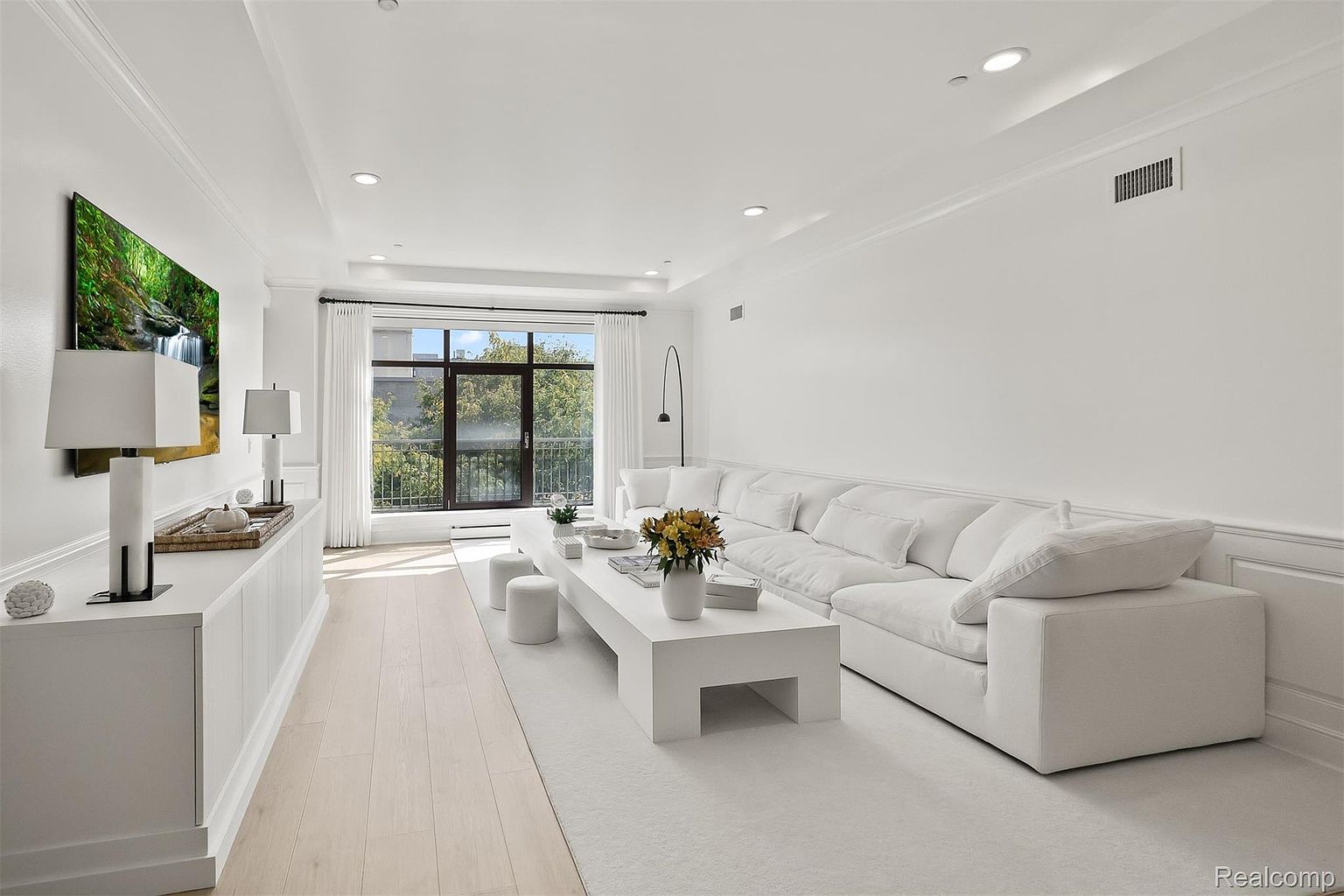 This is a bright and airy living room featuring a large white sectional sofa, a modern white coffee table, and a wall-mounted television displaying a nature scene. The room is bathed in natural light from a large window, complemented by recessed lighting. The overall aesthetic is minimalist and clean, creating a serene and inviting atmosphere.