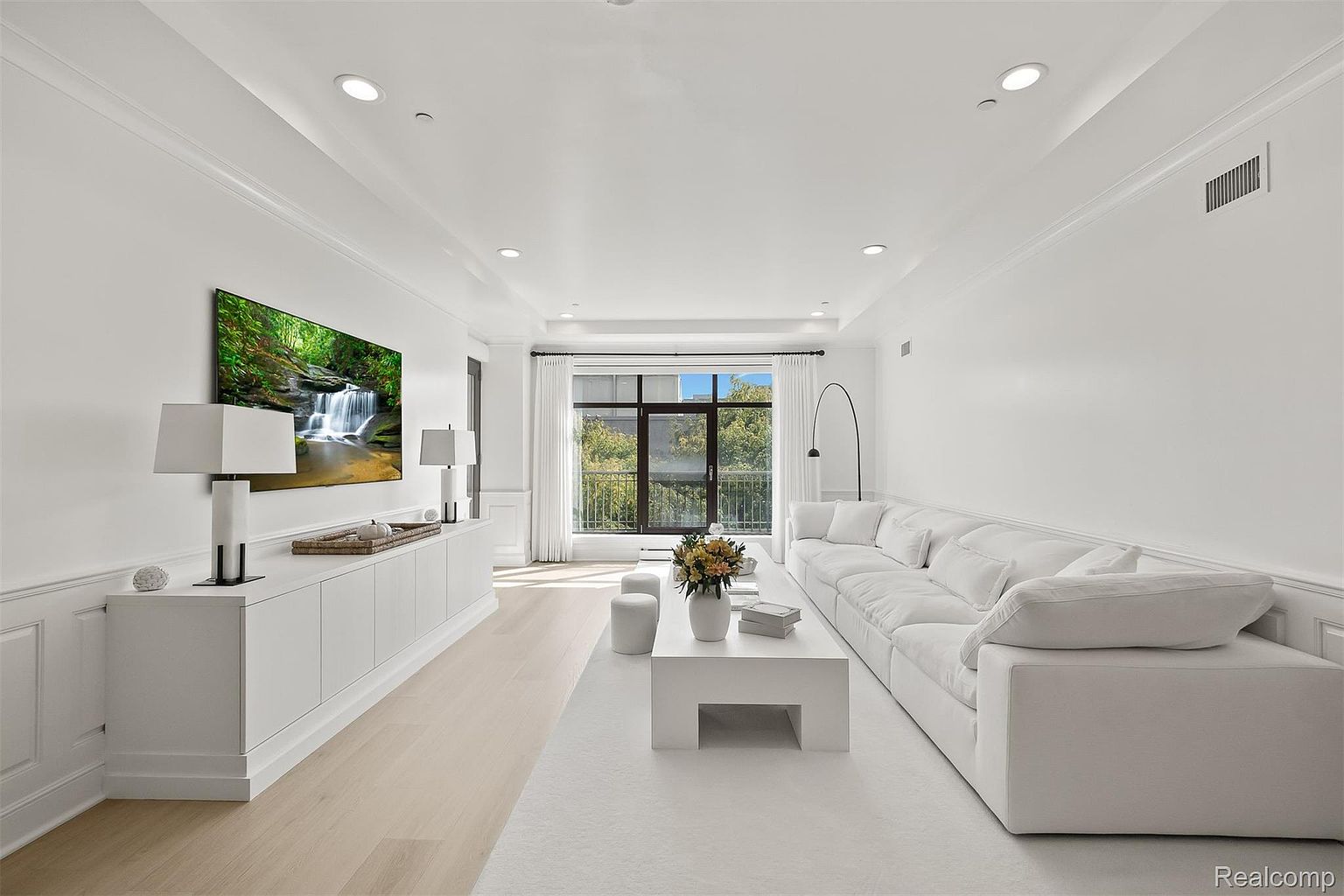 This is a bright and spacious living room featuring a large white sectional sofa, a modern white coffee table, and a long white media console with a large screen television mounted above. The room is decorated in a minimalist style with white walls, light wood flooring, and large windows that provide ample natural light. The overall impression is clean, modern, and inviting.