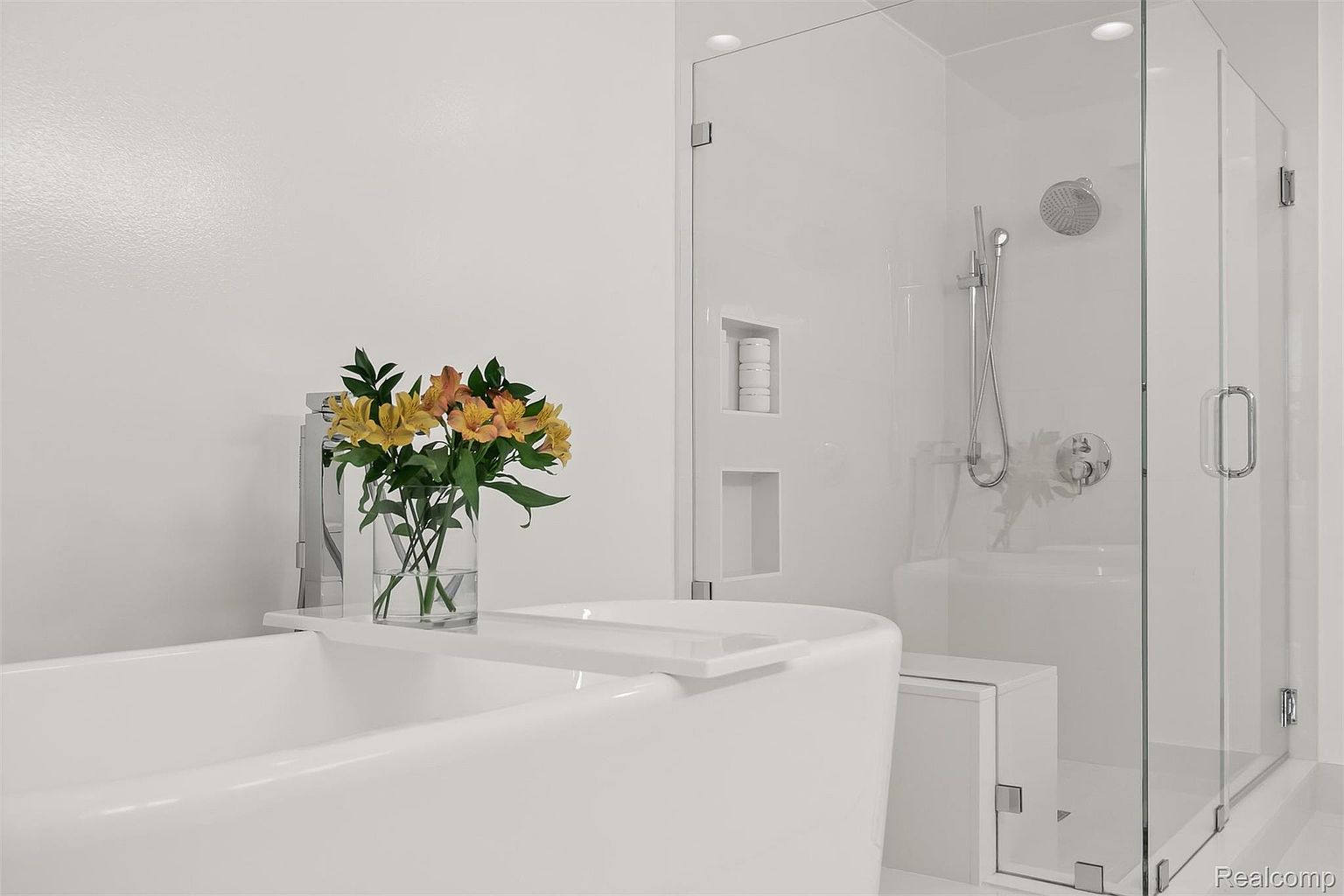 This is a bright and modern primary bathroom featuring a freestanding white bathtub with a clear glass shower enclosure. The bathroom has a clean, minimalist design with white walls and fixtures. A vase of flowers adds a touch of color and elegance to the space.