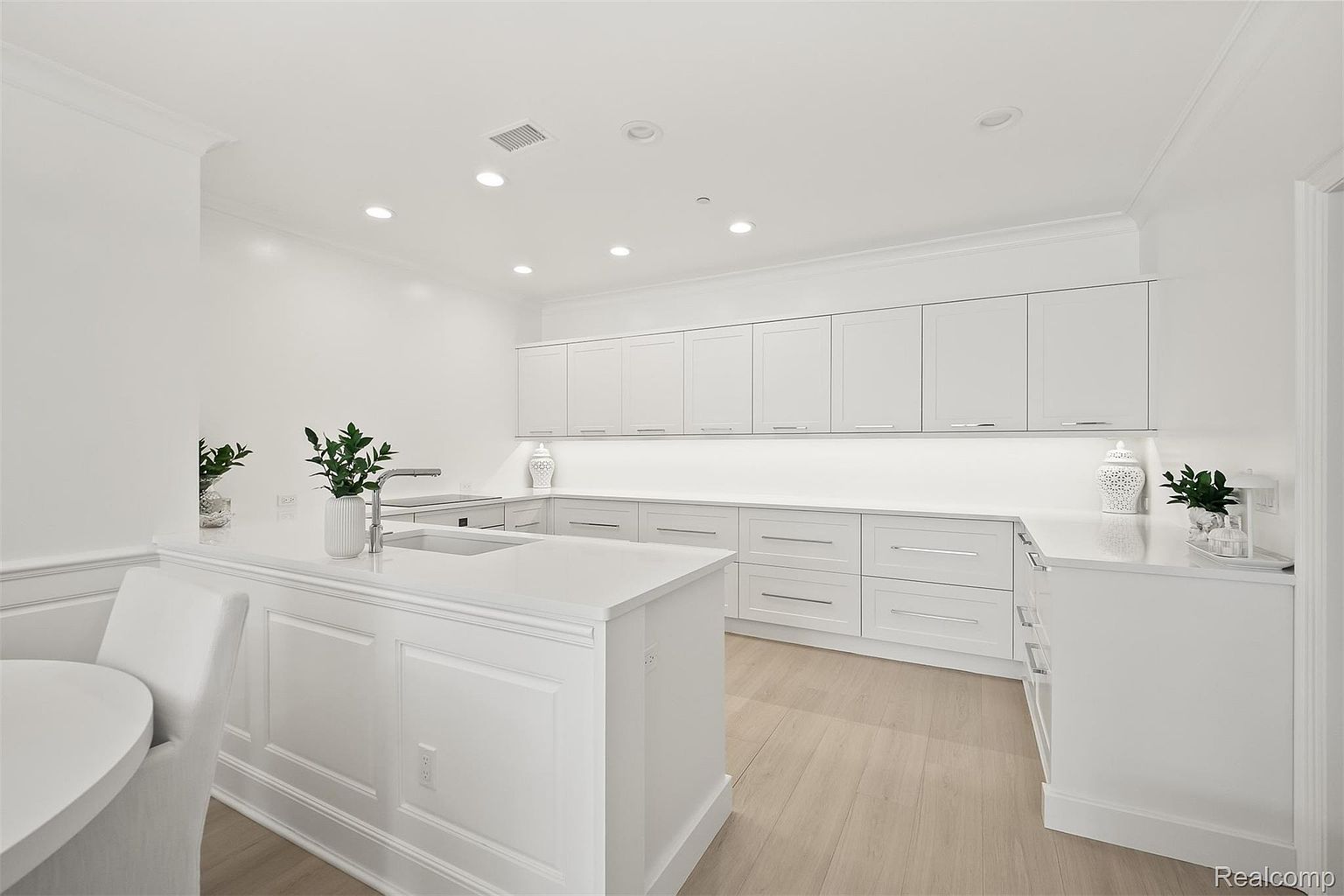 This is a bright, all-white kitchen featuring sleek cabinetry, a large island with a sink, and light wood flooring. The kitchen is well-lit with recessed lighting and under-cabinet lighting, creating a clean and modern aesthetic. The perspective is from a wide angle, showcasing the entire kitchen space.
