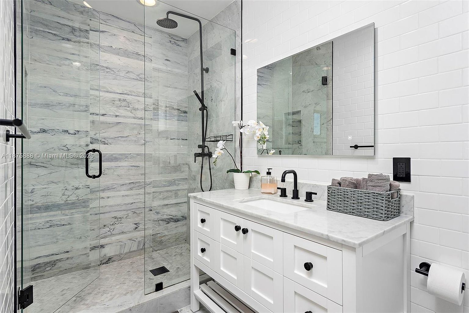 This is a primary bathroom featuring a glass-enclosed shower with marble-like tile and a black shower system. The vanity is white with a marble countertop, black hardware, and a modern faucet. A large mirror hangs above the vanity, reflecting the shower, and the walls are tiled in a classic white subway pattern, creating a clean and luxurious space.