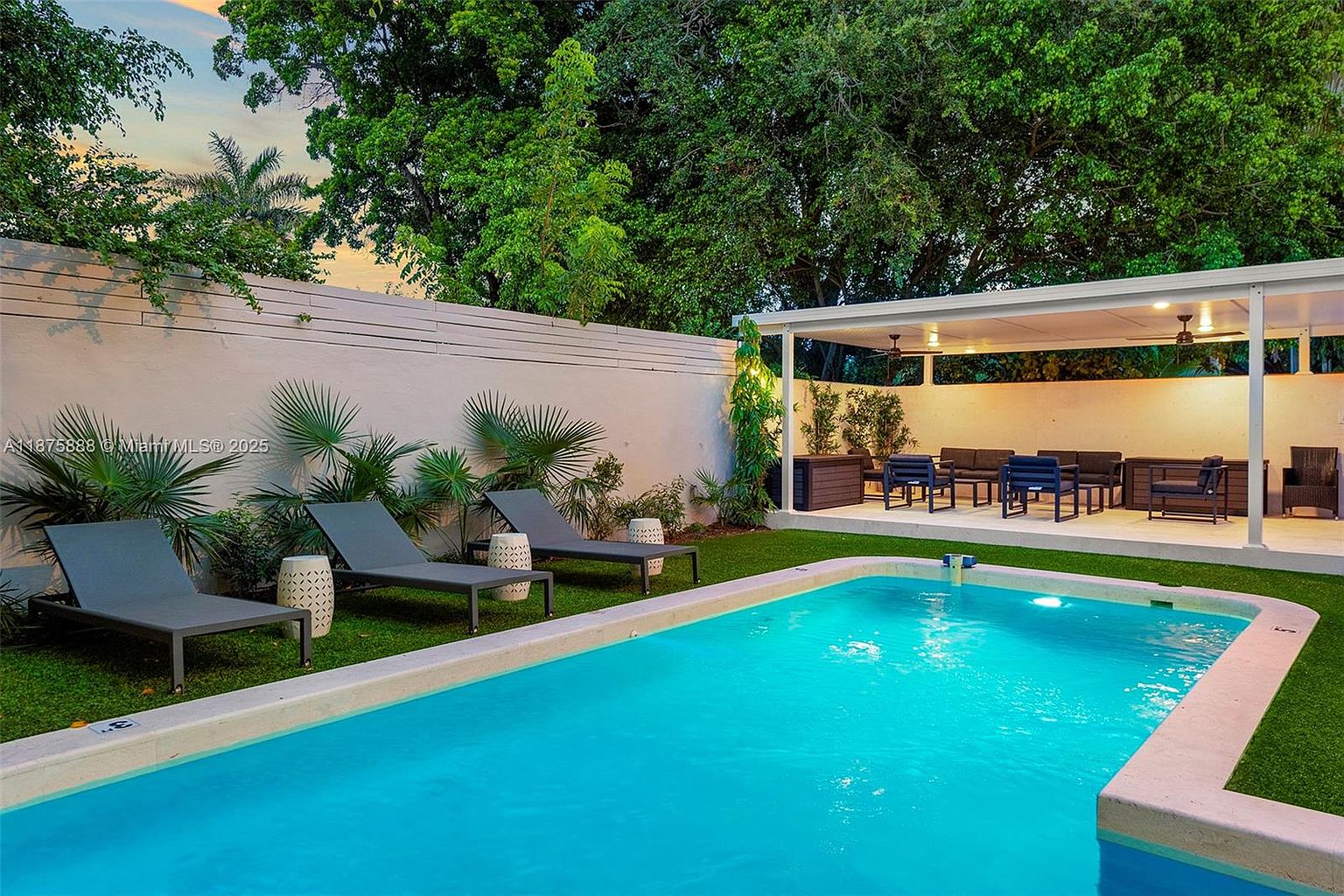This image showcases a luxurious backyard pool area, featuring a rectangular pool with clear blue water, surrounded by well-maintained green grass and lounge chairs. A covered patio with outdoor seating provides a comfortable space for relaxation and entertainment. The overall impression is one of a private and upscale outdoor oasis.