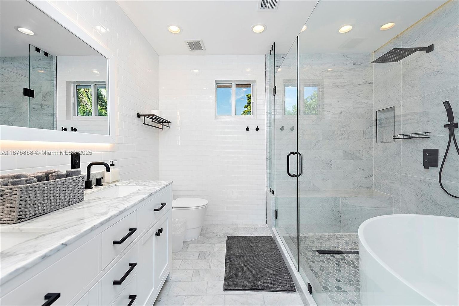 This is a bright and modern primary bathroom featuring white subway tile walls, marble flooring, and a glass-enclosed shower. The vanity has a white cabinet with a marble countertop and black hardware, complemented by a black faucet and accessories. The shower showcases marble walls, a built-in bench, and a rainfall showerhead, creating a luxurious and spa-like atmosphere.