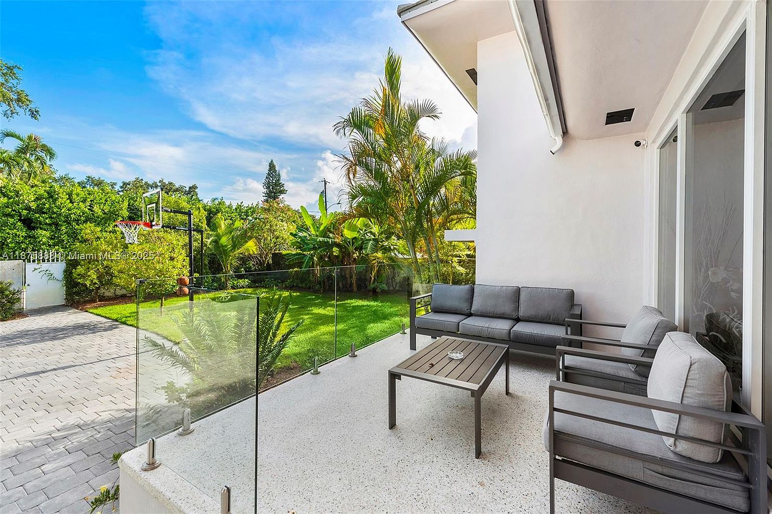 This image showcases a modern patio area with sleek outdoor furniture, including a gray sofa, two armchairs, and a coffee table. The patio features a light-colored, speckled concrete surface and a glass railing that offers an unobstructed view of the lush green lawn and landscaping. The exterior of the house is a clean white, contributing to the contemporary aesthetic.