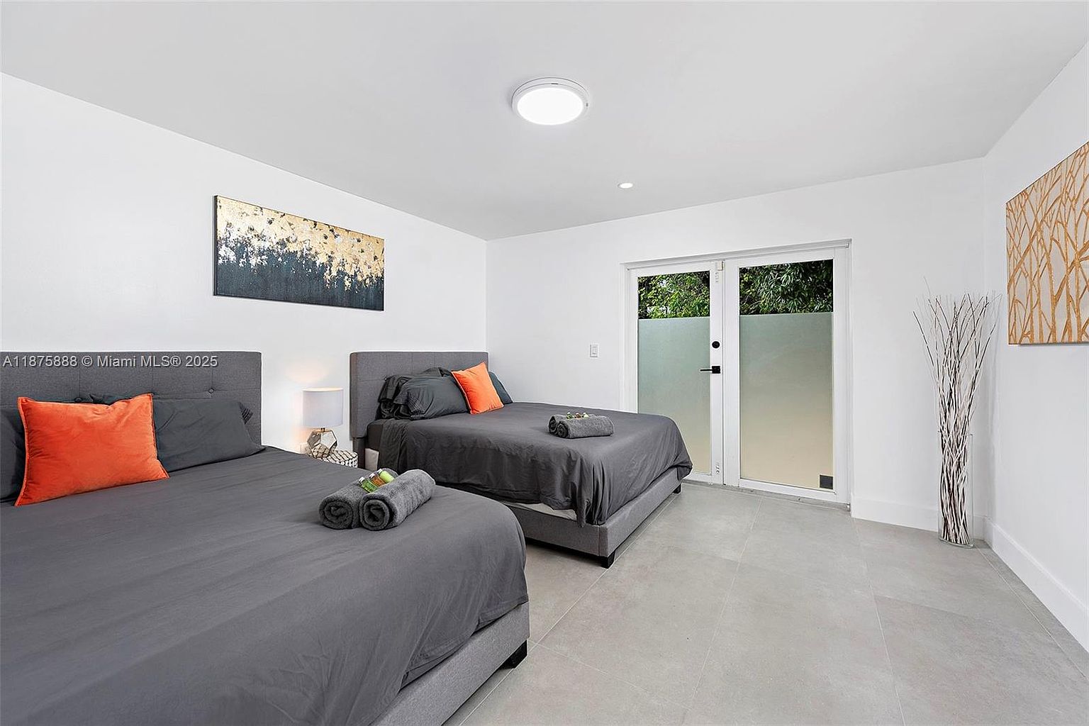 This is a bright and airy guest bedroom featuring two beds with gray linens and orange accent pillows. The room has white walls, light gray tile flooring, and modern artwork. A sliding glass door provides natural light and access to the outside, enhancing the room's appeal.