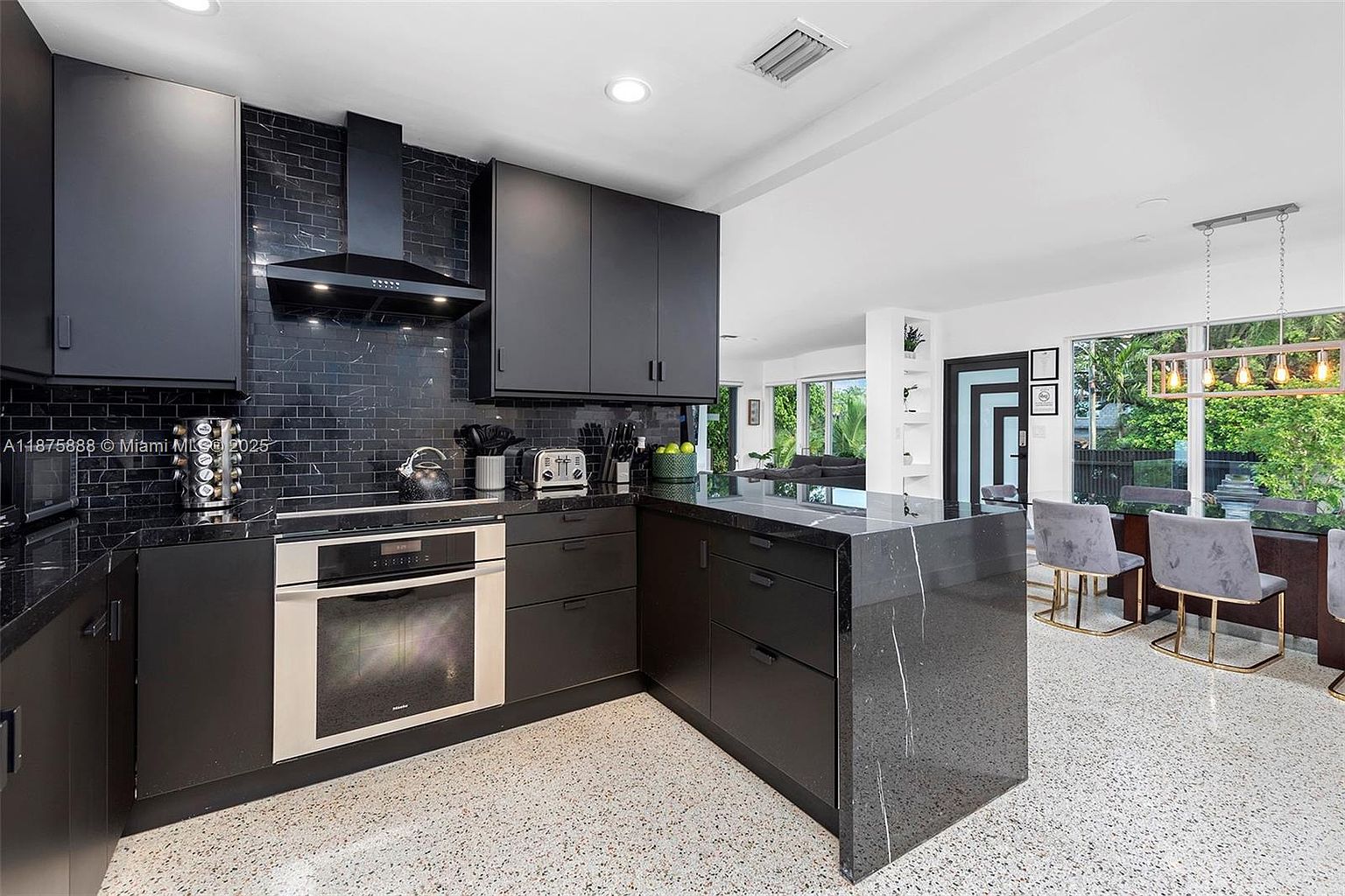 This is a modern kitchen with sleek, dark cabinetry and a contrasting light-colored terrazzo floor. The kitchen features stainless steel appliances, including a built-in oven, and a black marble countertop island. The open layout connects to a dining area with large windows, providing ample natural light and a view of the outdoors.