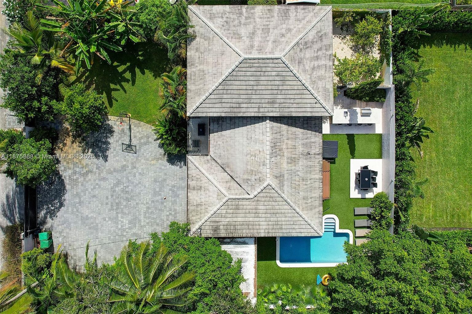 This aerial shot showcases a luxurious property featuring a large house with a gray tiled roof, a swimming pool surrounded by lush greenery, and an outdoor dining area with modern furniture. The property also includes a paved driveway with a basketball hoop and well-maintained lawns, creating an inviting and upscale atmosphere. The overall impression is one of privacy, elegance, and outdoor living.