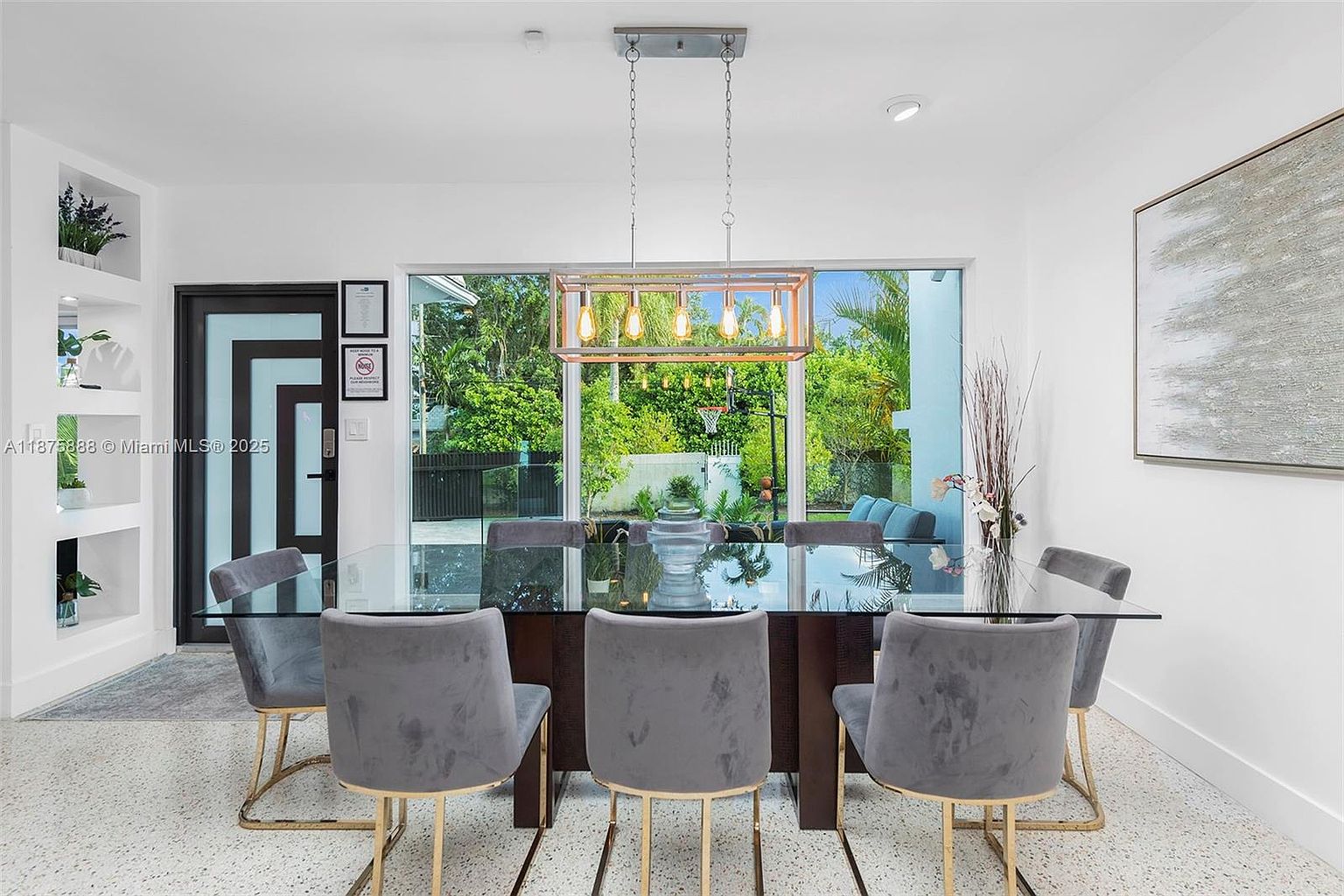 This is an interior shot of a modern dining room. The room features a glass-topped dining table with gray velvet chairs and gold legs, illuminated by a rectangular pendant light fixture. A large window provides a view of the outdoor greenery, and a piece of abstract art hangs on the wall, contributing to the room's contemporary aesthetic.