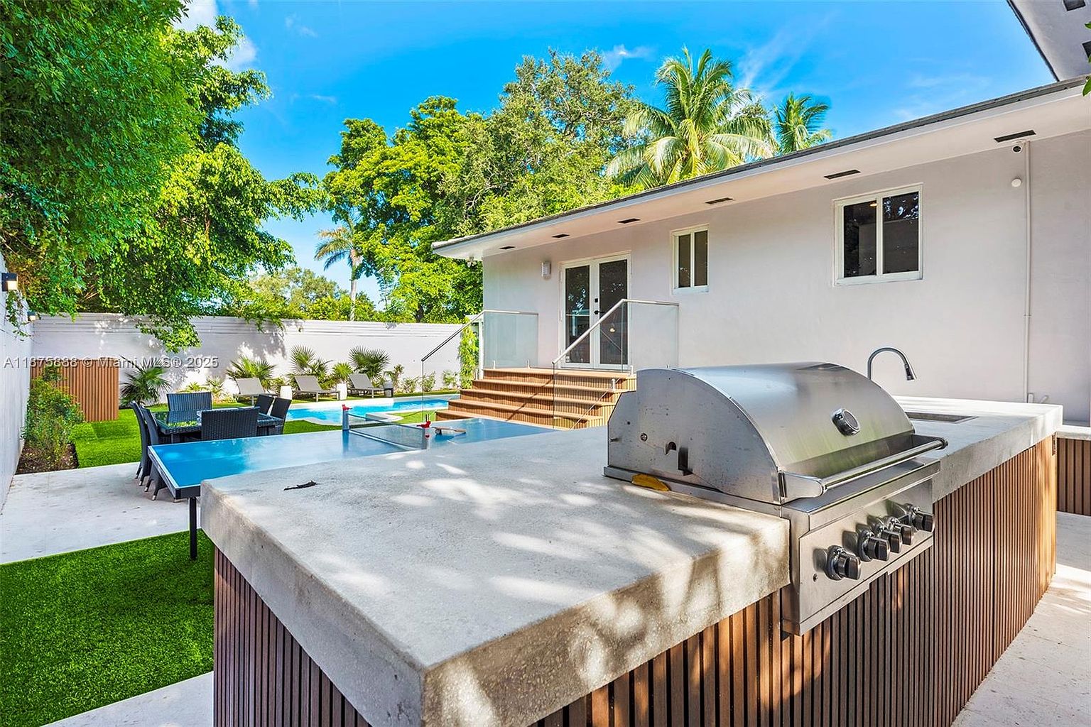 This image showcases a beautifully landscaped backyard featuring a modern outdoor kitchen with a built-in grill and concrete countertop. The space includes a dining area, a ping pong table, and a swimming pool, all surrounded by lush greenery and a white privacy fence. The overall impression is one of luxury and relaxation, perfect for outdoor entertaining.