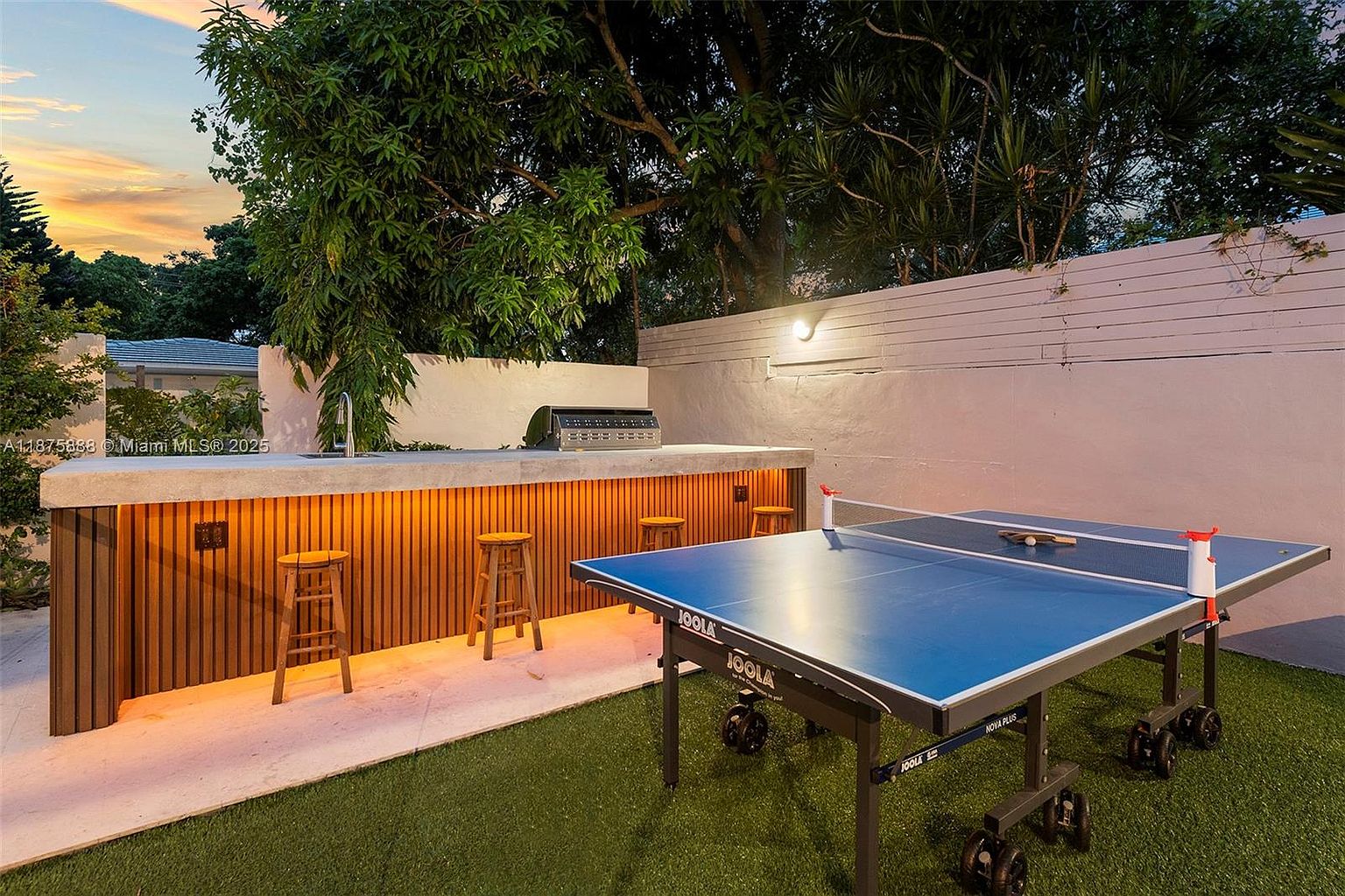 This image showcases a well-maintained backyard area, featuring an outdoor kitchen with a built-in grill and bar seating. A ping pong table sits on the artificial turf, suggesting a recreational space. The area is enclosed by a white wall, and mature trees provide shade and privacy, creating an inviting outdoor living space.