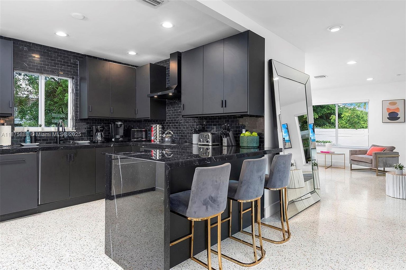 This is a modern kitchen with sleek, dark cabinetry and black countertops. The kitchen features a breakfast bar with three gray velvet bar stools with gold legs. A large mirror leans against the wall, reflecting the adjacent living area, and the overall design is contemporary and stylish.