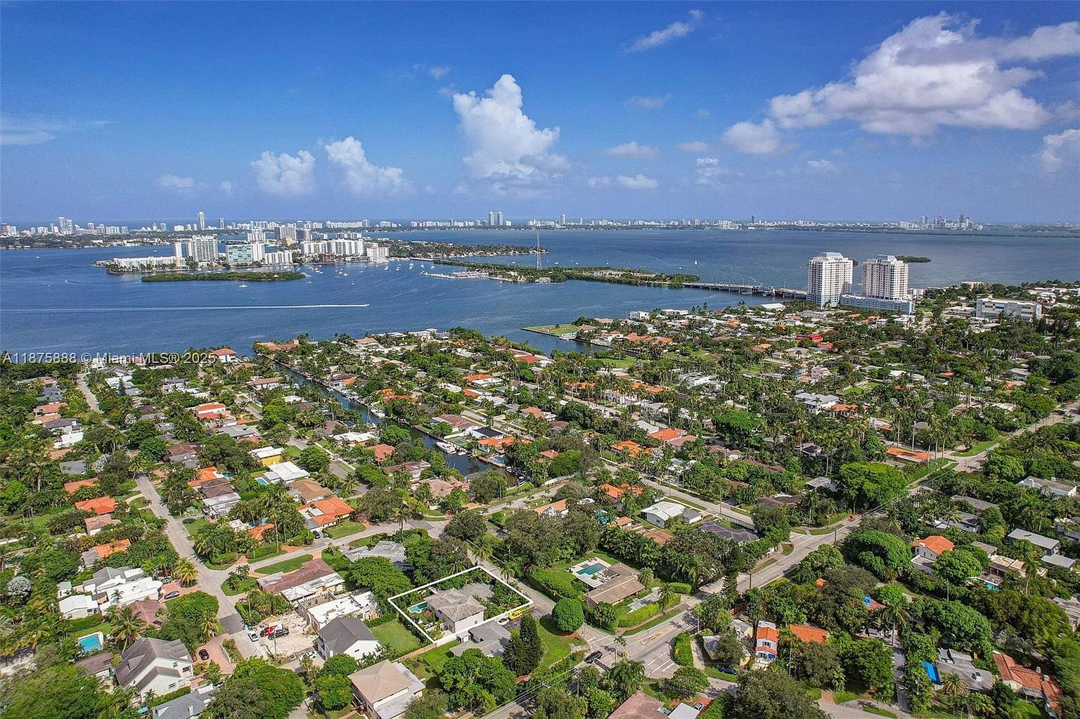This aerial view showcases a residential area with numerous houses, lush greenery, and waterways. The houses feature a mix of architectural styles, and many have pools. In the background, a cityscape is visible across the water, creating a scenic backdrop.