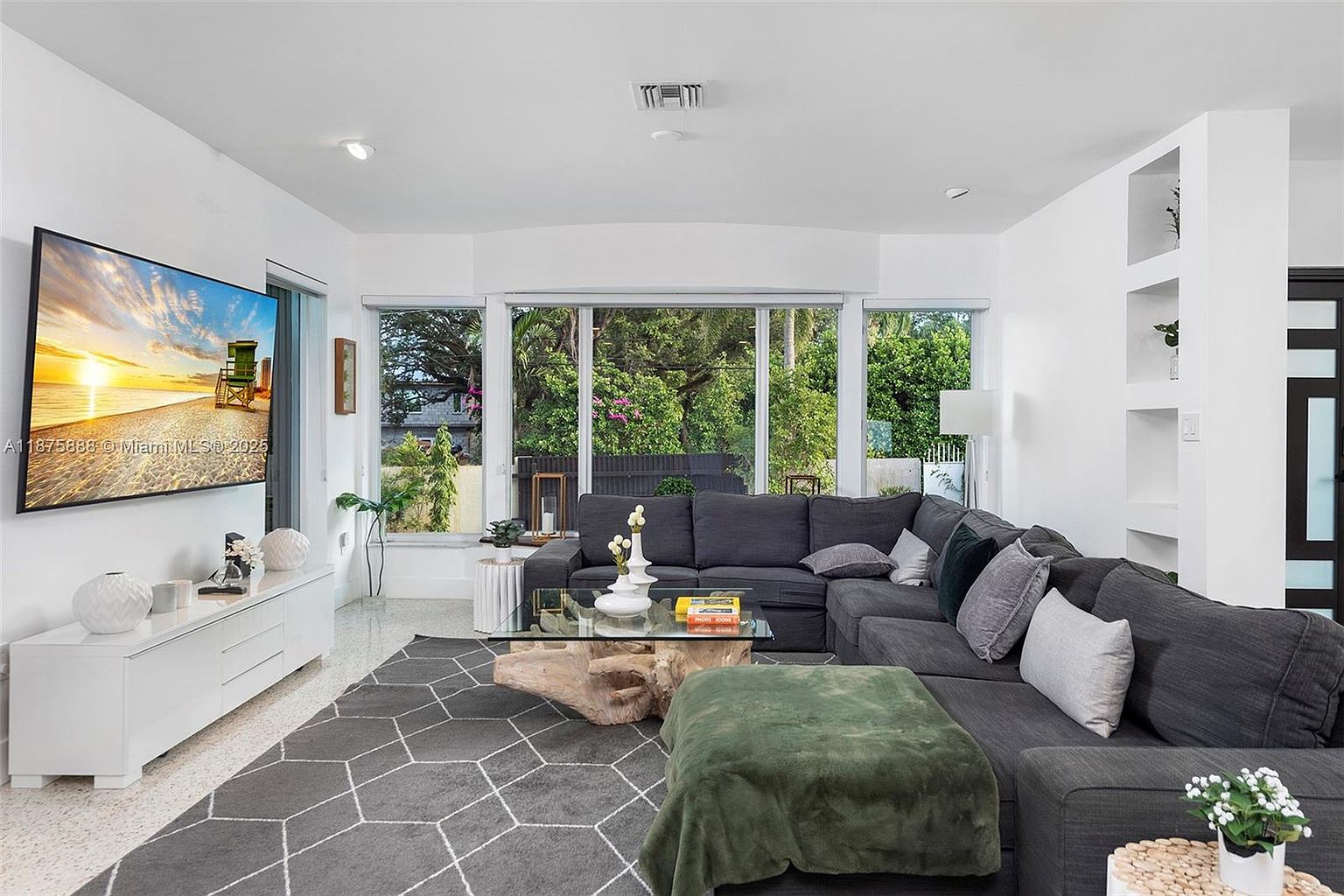 This is a spacious living room featuring a large, comfortable gray sectional sofa adorned with decorative pillows. A unique glass-topped coffee table with a natural wood base sits atop a geometric patterned gray rug. Large windows provide ample natural light and views of the lush greenery outside, while a modern entertainment center houses a large flat-screen TV.