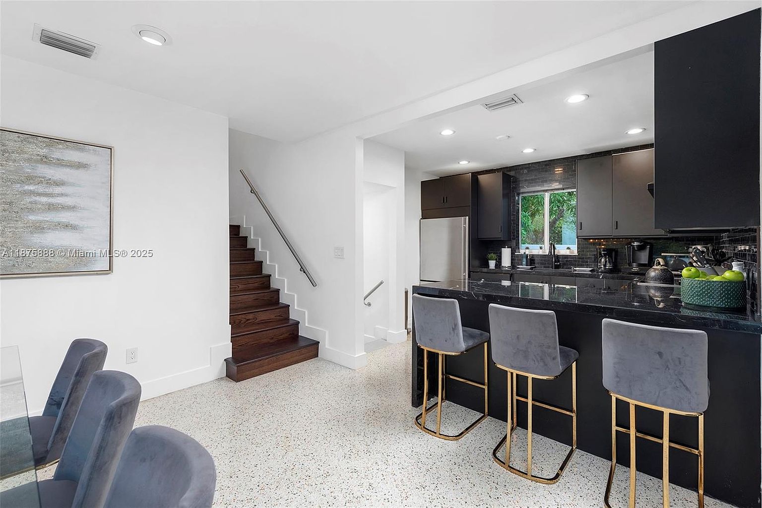 This interior shot showcases a modern kitchen and dining area. The kitchen features dark cabinetry, stainless steel appliances, and a sleek black countertop with bar seating. The dining area includes a glass-top table with gray upholstered chairs, and a staircase leads to another level, creating a stylish and functional living space.
