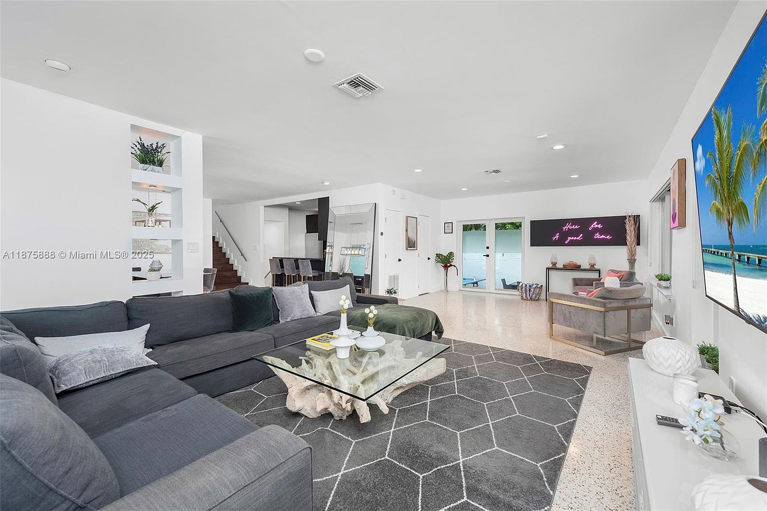 This is a spacious and modern living room featuring a large gray sectional sofa, a unique glass-topped coffee table with a driftwood base, and a gray geometric patterned rug. The room is well-lit with recessed lighting and natural light from the sliding glass doors, and it includes a large television and a decorative neon sign, creating a stylish and comfortable atmosphere. The open floor plan leads to other areas of the house, enhancing the sense of space.