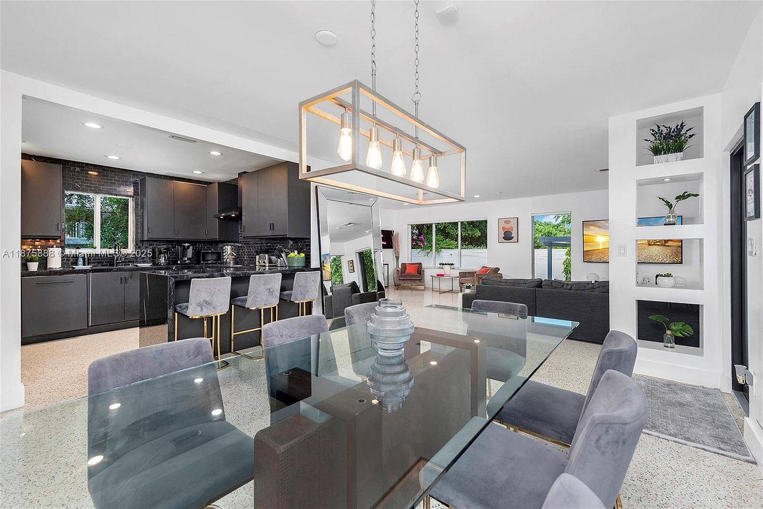 This interior shot showcases a modern dining area adjacent to a kitchen and living space. A glass-topped dining table with gray upholstered chairs sits beneath a contemporary rectangular chandelier. The kitchen features dark cabinetry and a breakfast bar, while the living area is visible in the background, creating an open and inviting atmosphere.