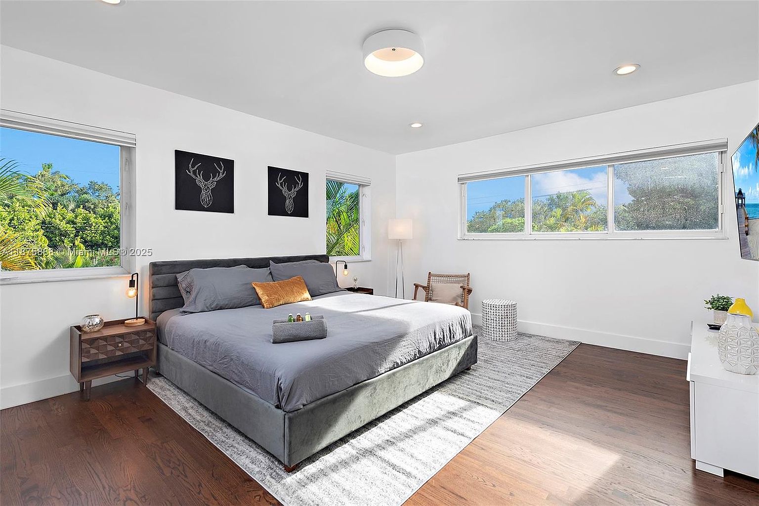 This is a well-lit primary bedroom featuring a large bed with gray bedding and a gold accent pillow. Two black and white deer head art pieces hang above the bed. The room has hardwood floors, a gray rug, and large windows that provide natural light and views of the outside greenery, creating a serene and inviting atmosphere.