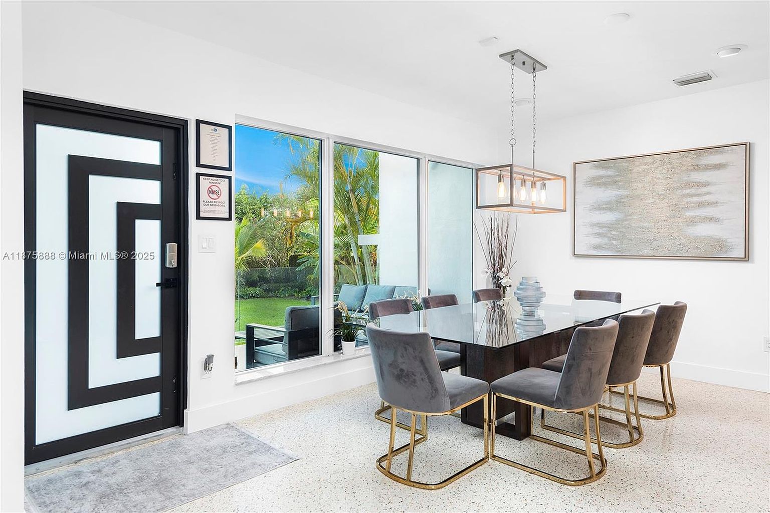 This is a well-lit dining room featuring a modern aesthetic. A glass-topped dining table is surrounded by six gray velvet chairs with gold legs, illuminated by a contemporary chandelier. A large window offers a view of the outdoor greenery, and an abstract painting hangs on the wall, adding a touch of sophistication.