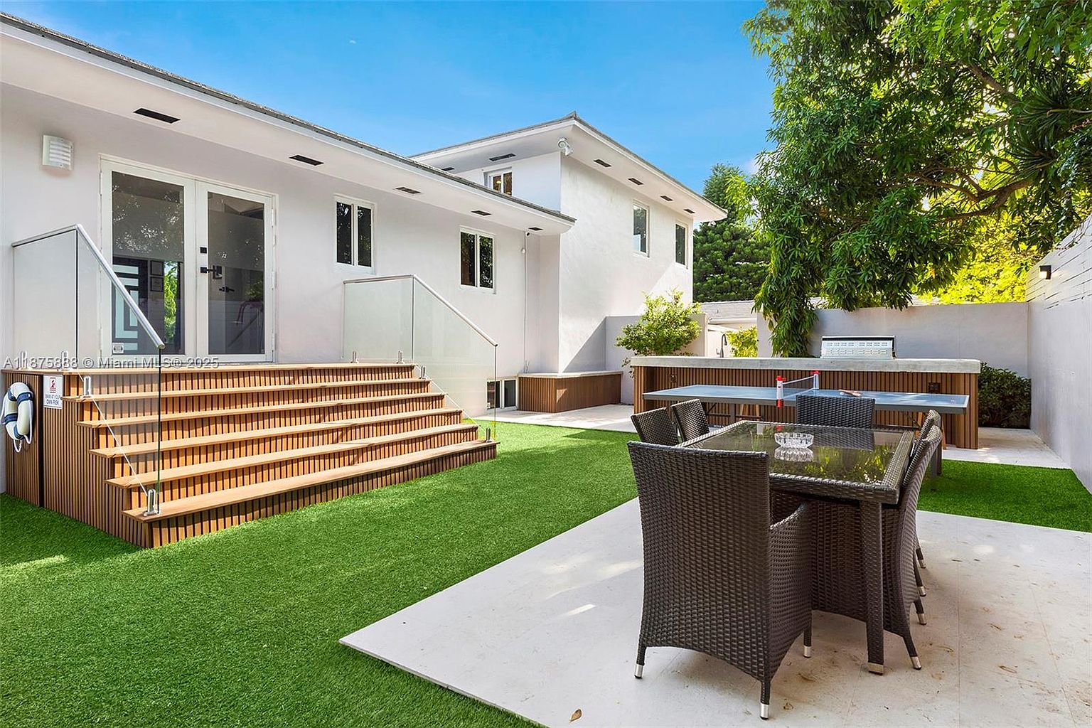 This image showcases a well-maintained backyard featuring a modern home with a multi-level wooden deck leading to a manicured lawn. An outdoor dining area with wicker furniture and a built-in grill area provide ample space for entertaining. The scene is bathed in natural light, creating a serene and inviting atmosphere.
