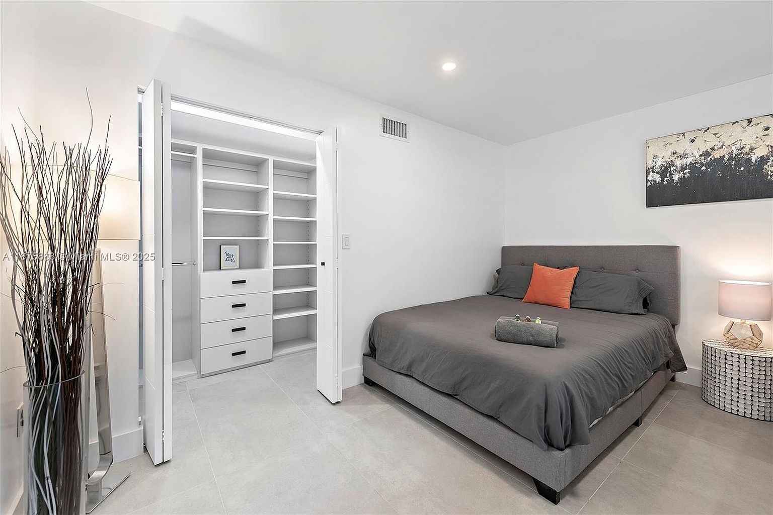 This is a well-lit primary bedroom featuring a gray upholstered bed with an orange accent pillow, a modern nightstand with a lamp, and a piece of abstract art on the wall. The room has a walk-in closet with white shelving and drawers, contributing to a clean and organized aesthetic. The flooring is light gray tile, and the walls are painted white, creating a bright and airy atmosphere.