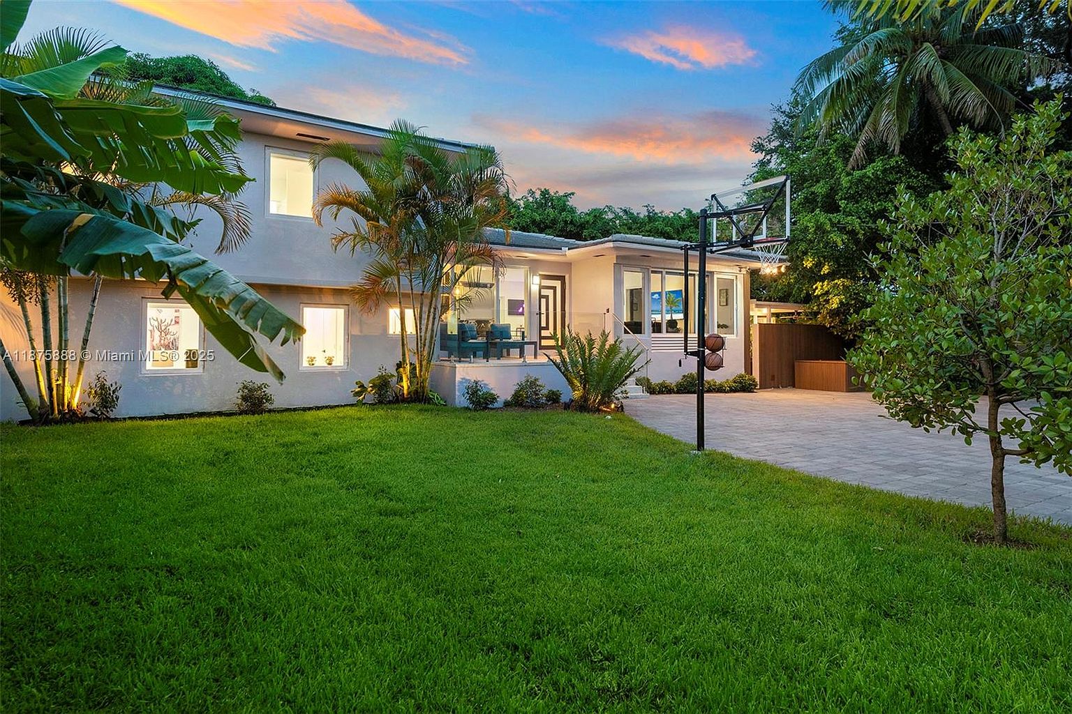 This image showcases the front exterior of a modern two-story home with a well-manicured lawn. The house features a light-colored facade, large windows, and a contemporary design with palm trees and other lush greenery surrounding the property. A basketball hoop stands on the paved driveway, adding a touch of recreational appeal.