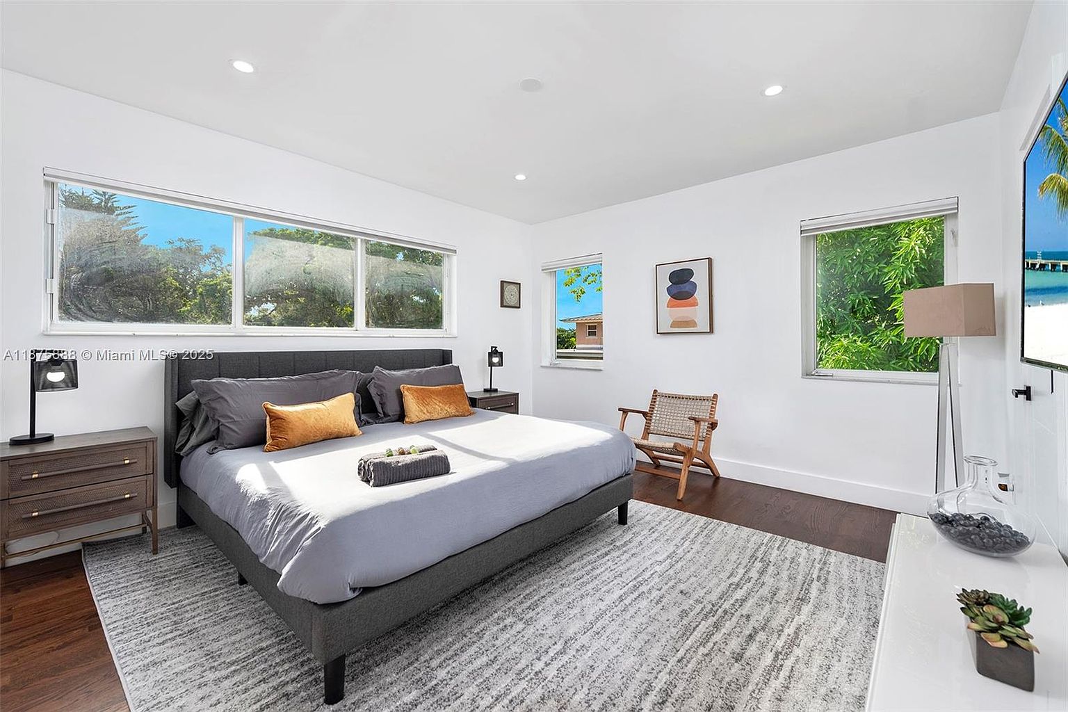 This is a well-lit primary bedroom featuring a gray upholstered bed with gray and gold pillows, a gray and white patterned rug, and dark hardwood floors. The room has white walls and multiple windows providing natural light, complemented by modern artwork and minimalist decor. The overall impression is clean, modern, and inviting.