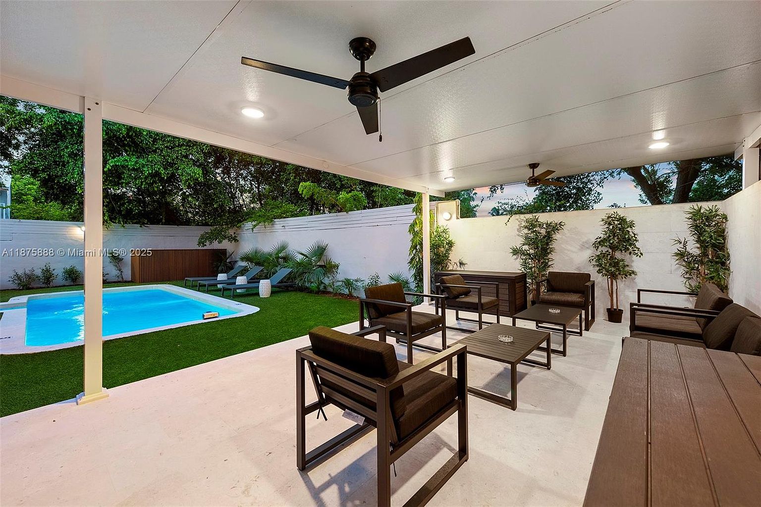 This image showcases a stylish outdoor patio area with modern furniture, including chairs, a coffee table, and a long bench. The patio is covered, featuring ceiling fans and recessed lighting, and it overlooks a well-maintained lawn and a swimming pool. The design emphasizes a comfortable and inviting outdoor living space, perfect for relaxation and entertainment.