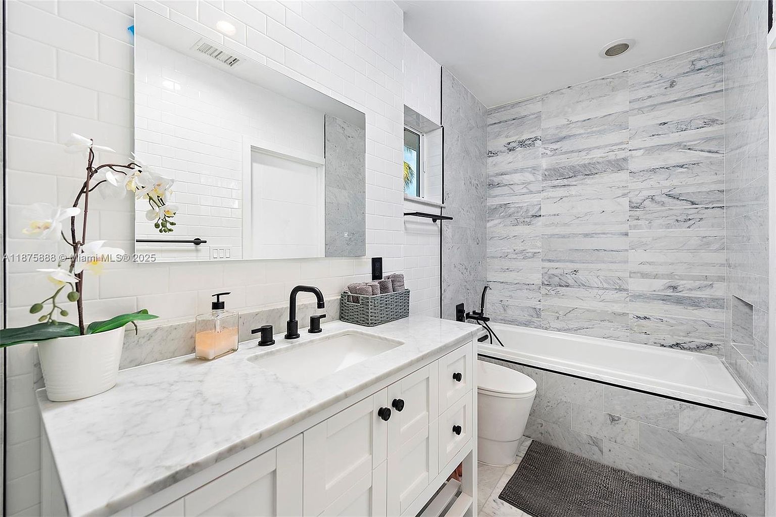 This is a primary bathroom featuring a white marble countertop vanity with black fixtures and white cabinetry. The walls are tiled with white subway tiles and marble-like tiles around the bathtub. The bathroom has a clean and modern aesthetic, with a focus on bright, neutral colors and high-end finishes.