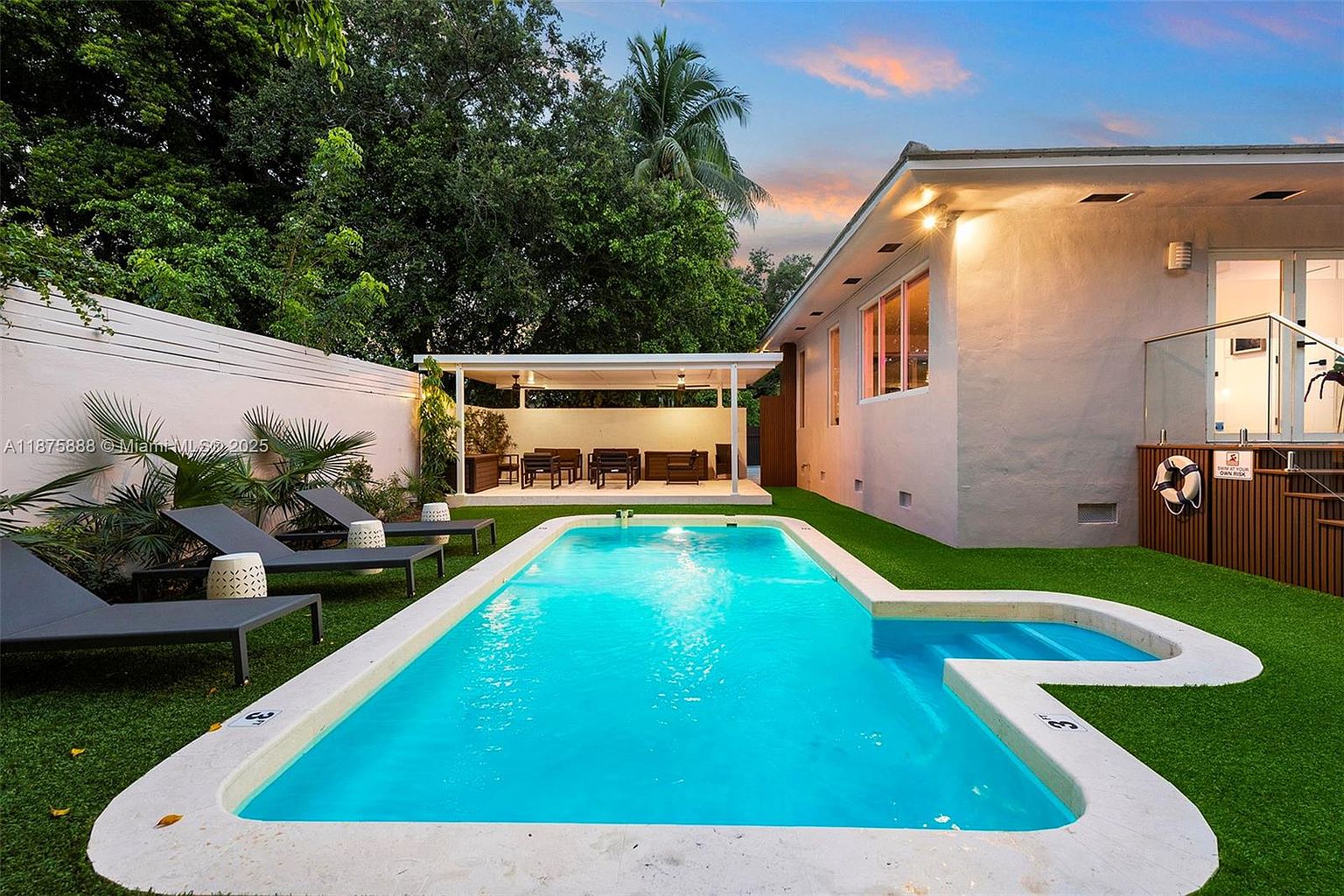 This image showcases a beautifully maintained backyard featuring a rectangular swimming pool with a shallow end, surrounded by lush green artificial turf. Lounge chairs are placed near the pool, and a covered patio area with outdoor seating is visible in the background. The property is enclosed by a white fence, providing privacy, and the overall impression is one of relaxation and luxury.