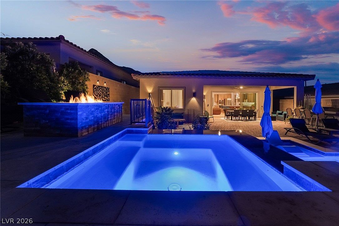 This twilight shot captures a luxurious backyard oasis featuring a glowing, blue-lit swimming pool in the foreground. To the left, a stone-clad fire feature provides a warm contrast, while the covered patio area in the background is furnished with an outdoor dining set and lounge chairs. The scene is set against a vibrant sunset sky, creating an inviting and sophisticated atmosphere for evening relaxation.
