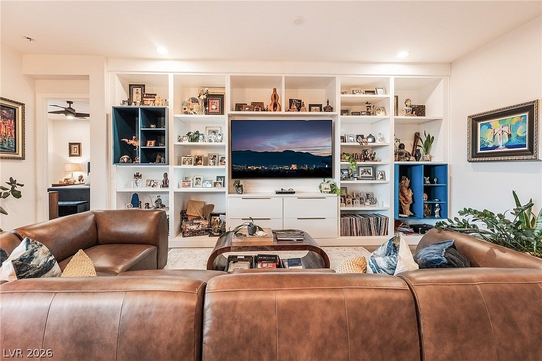 This inviting living room features a large, brown leather sectional sofa arranged around a dark wood coffee table. The focal point is a massive, custom-built white shelving unit that spans the wall, housing a television, decorative items, and books, with two distinct blue-accented cabinet sections. The space is well-lit with recessed lighting and offers a warm, comfortable atmosphere suitable for family gatherings.