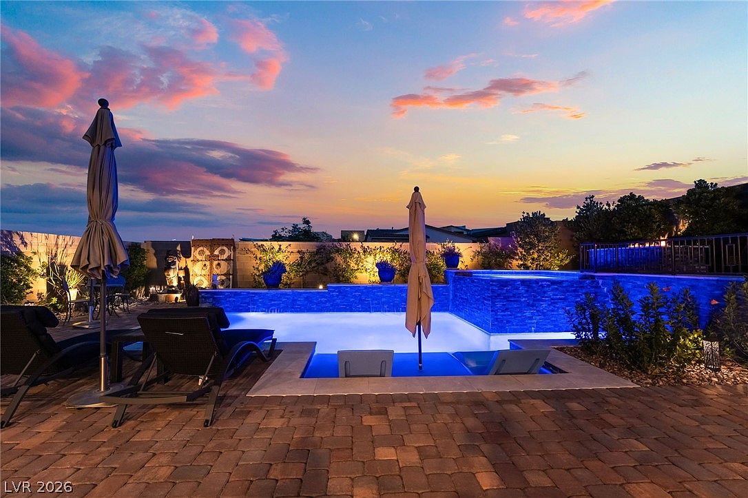 This backyard oasis features a modern swimming pool with integrated blue LED lighting and a raised spa section. The area is furnished with lounge chairs and patio umbrellas, set against a backdrop of a vibrant sunset sky. The space is enclosed by a stone wall with decorative metal accents and landscaping, creating a private and inviting atmosphere for evening relaxation.
